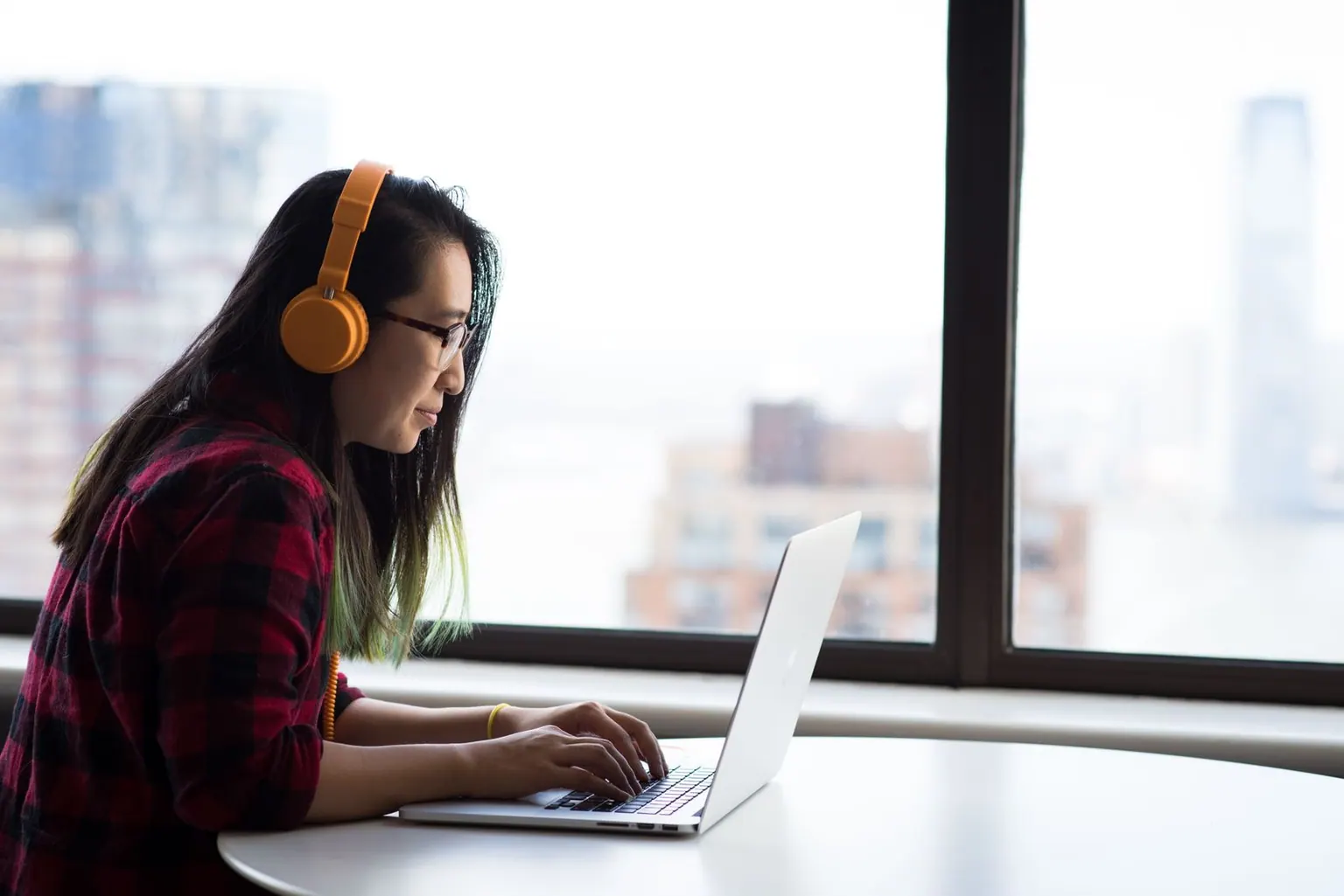 An Asian woman working remotely on a laptop with headphones beside a city window.