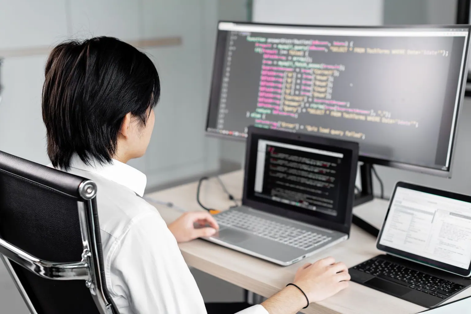 An Asian developer writing code across a laptop and a wide desktop monitor.
