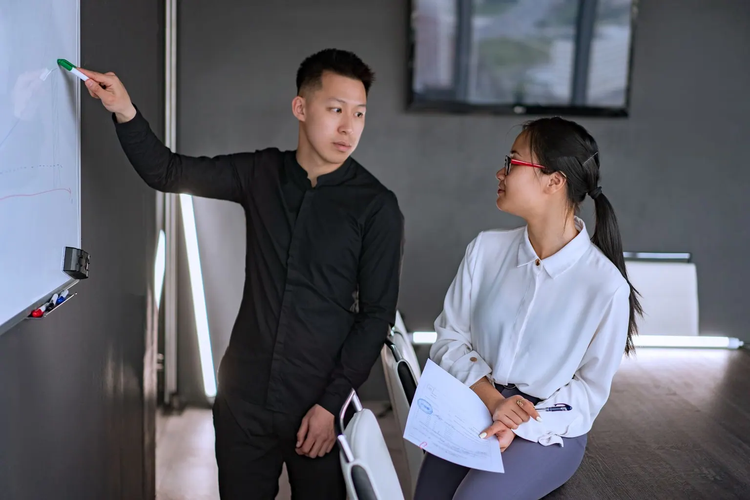 An Asian team lead mapping out delivery plans on a whiteboard with a colleague.