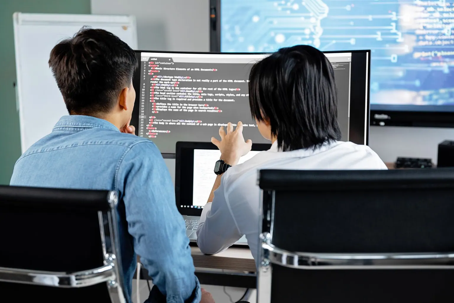 Two Asian software developers reviewing code together on a large monitor in an office.