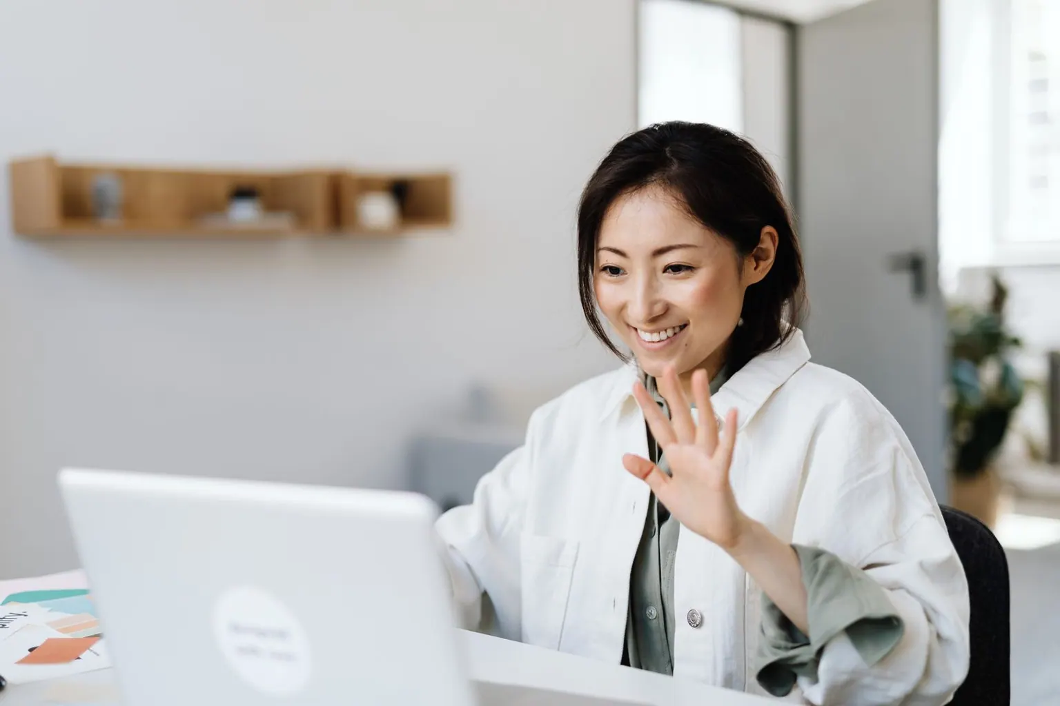 An Asian professional waving to her overseas team at the start of a video call.