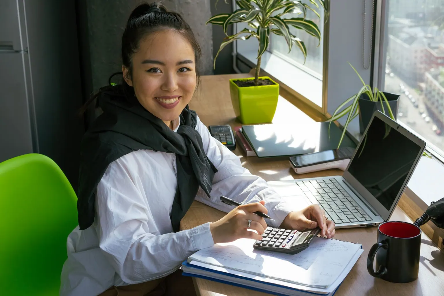 An Asian accountant working through figures with a calculator beside a bright office window.